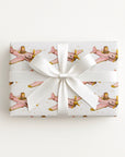 A rectangular gift box wrapped in ivory wrapping paper with a repeating pattern of vintage pink airplanes with shark mouth noses and gold accents, tied with a wide white satin ribbon and bow, on a plain white background.