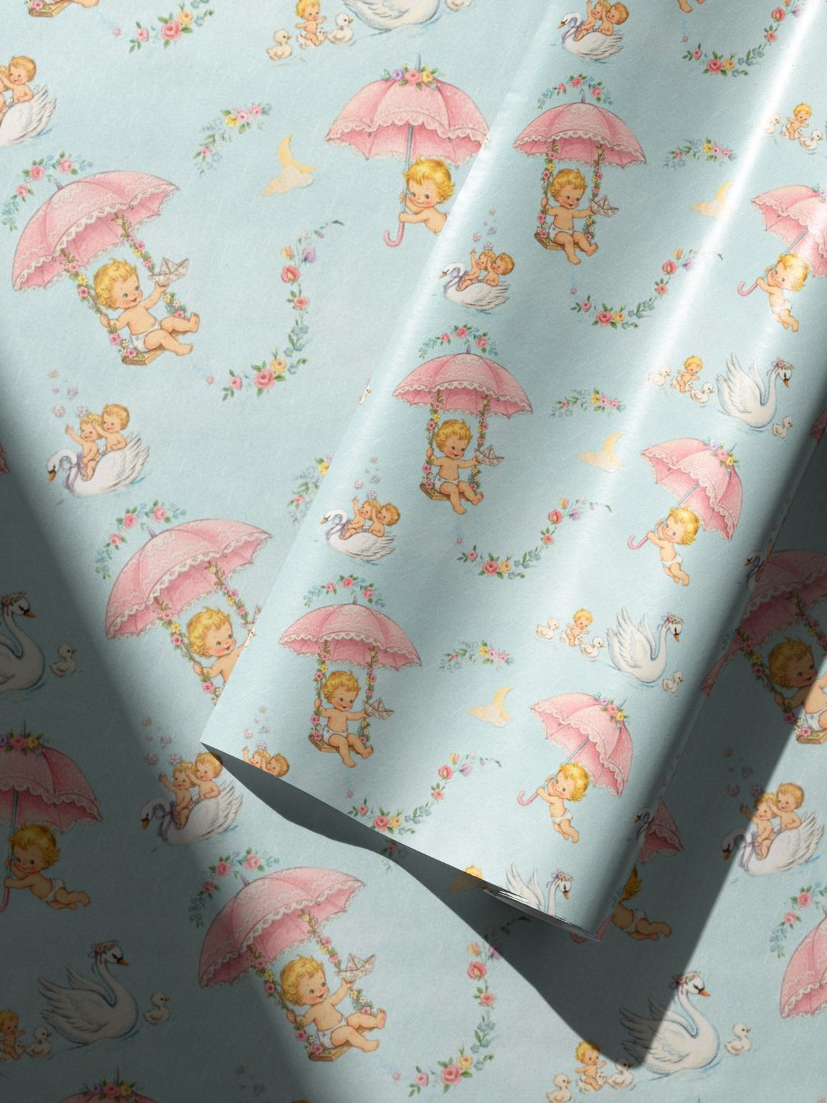 Light blue wrapping paper with a repeating pattern of darling babies sitting under pink umbrellas, swans, and small floral wreaths, shown partially rolled with soft shadows across the surface.
