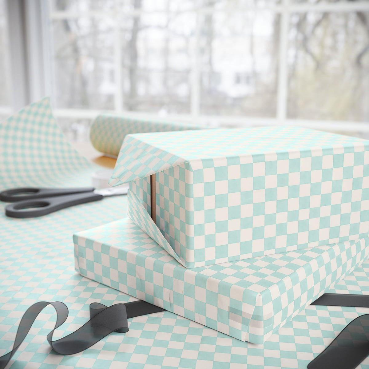 Light aqua and soft white checkered wrapping paper shown on gift boxes and a table, with scissors and ribbon nearby in front of a softly blurred window.