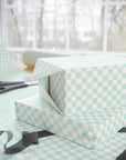 Light aqua and soft white checkered wrapping paper shown on gift boxes and a table, with scissors and ribbon nearby in front of a softly blurred window.