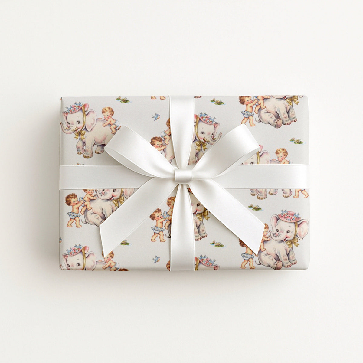 A rectangular gift box wrapped in ivory wrapping paper with a vintage-style illustration of elephants and babies, tied with a white satin ribbon and bow, photographed from above on a light gray background.