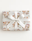A rectangular gift box wrapped in ivory wrapping paper with a vintage-style illustration of elephants and babies, tied with a white satin ribbon and bow, photographed from above on a light gray background.