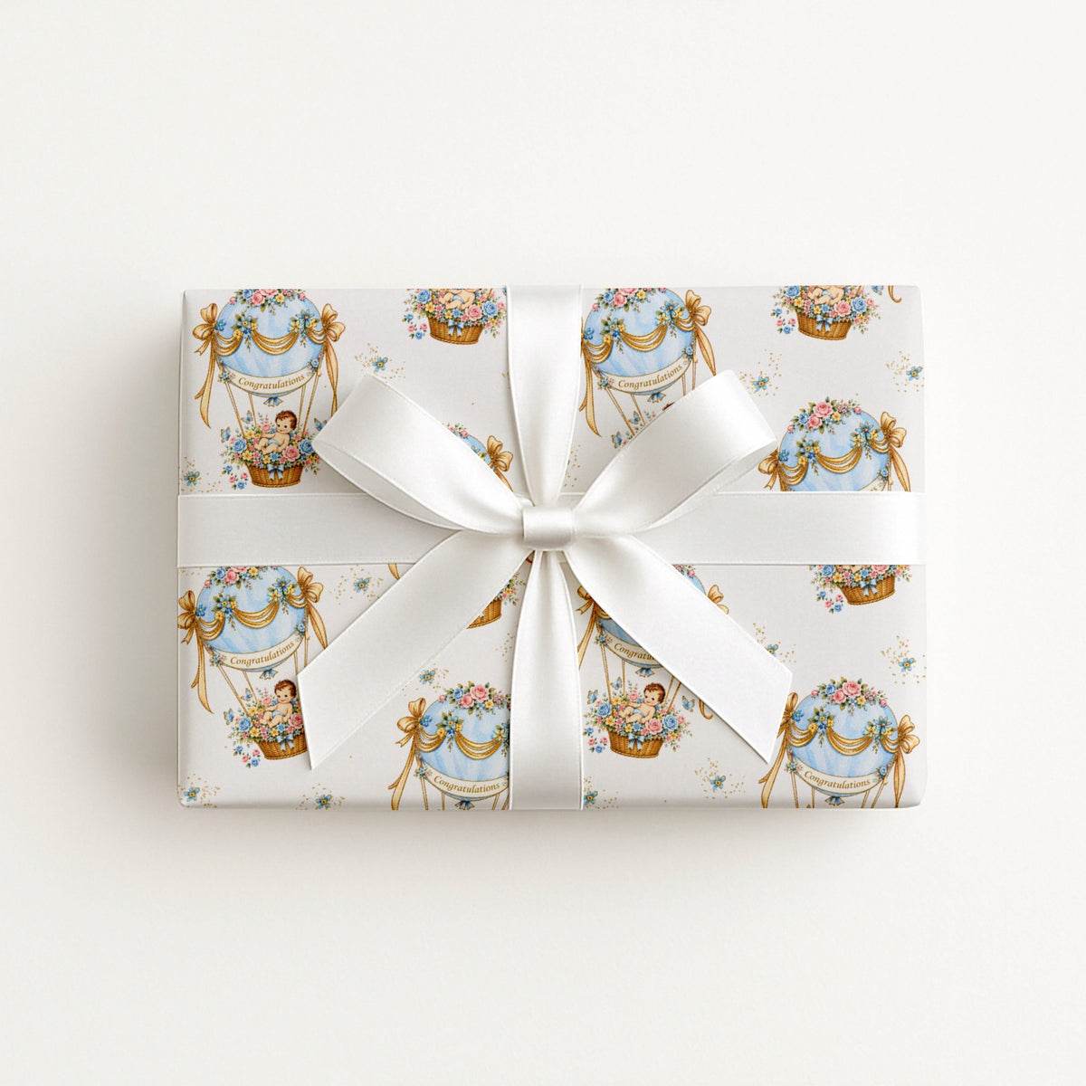 A rectangular gift box wrapped in cream baby shower wrapping paper with a repeating pastel illustration pattern of ornate hot air balloons and ribbon motifs, tied with a wide white satin bow in the center, photographed against a clean white background.