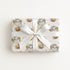 A rectangular gift box wrapped in cream baby shower wrapping paper with a repeating pastel illustration pattern of ornate hot air balloons and ribbon motifs, tied with a wide white satin bow in the center, photographed against a clean white background.