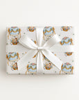 A rectangular gift box wrapped in cream baby shower wrapping paper with a repeating pastel illustration pattern of ornate hot air balloons and ribbon motifs, tied with a wide white satin bow in the center, photographed against a clean white background.