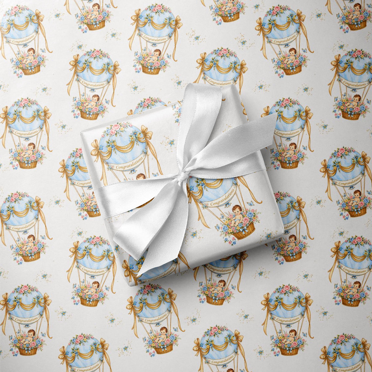 A wrapped baby boy gift box tied with a white satin ribbon, placed on top of matching wrapping paper printed with a repeating vintage hot air balloon motif in soft blue, gold, and pink on a white background.