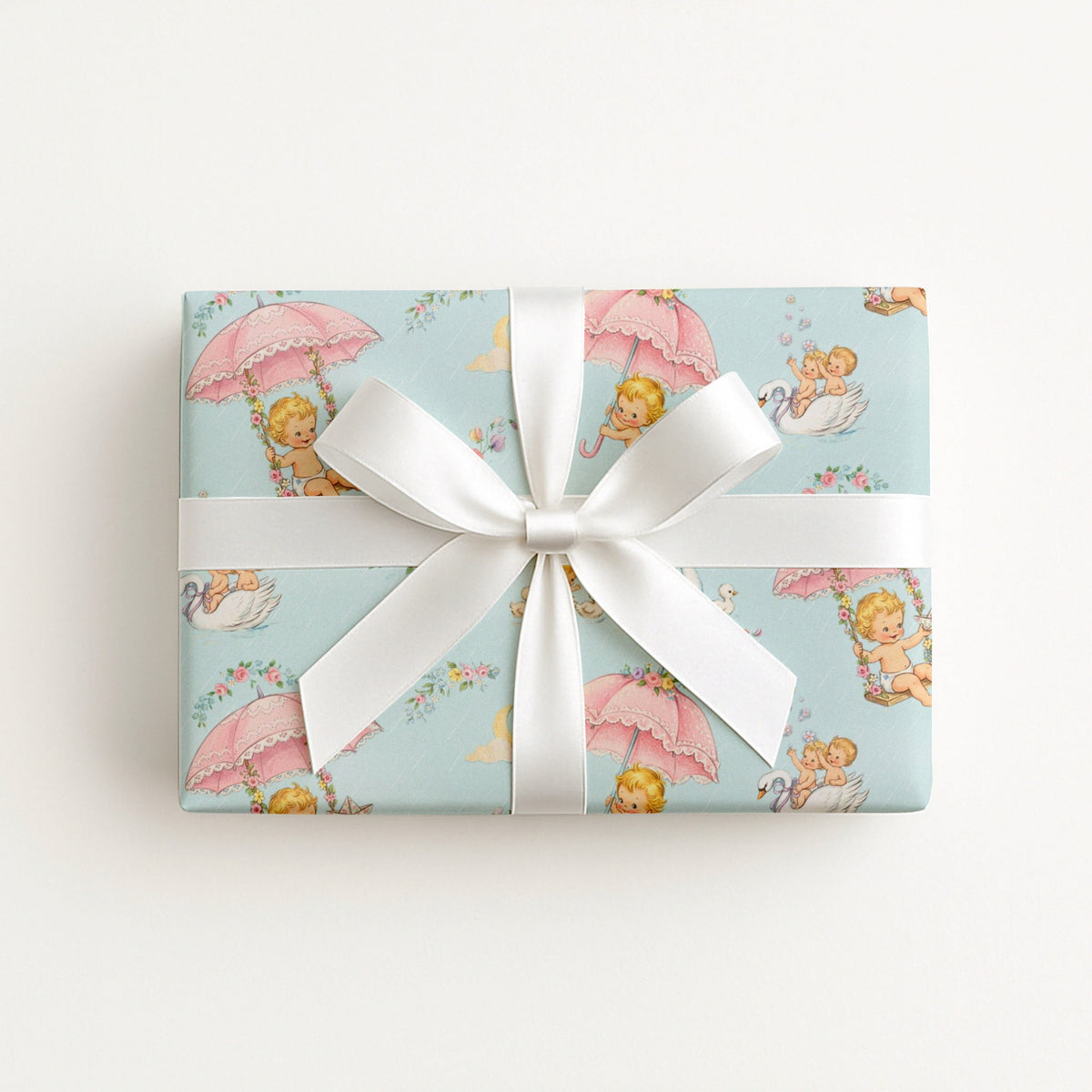 A rectangular gift box wrapped in vintage-inspired baby shower wrapping paper featuring a repeating 1950s storybook pattern of baby boys and girls floating on pink lace umbrellas and wooden swings against a pastel blue background with white swans.
