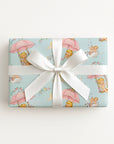 A rectangular gift box wrapped in vintage-inspired baby shower wrapping paper featuring a repeating 1950s storybook pattern of baby boys and girls floating on pink lace umbrellas and wooden swings against a pastel blue background with white swans.