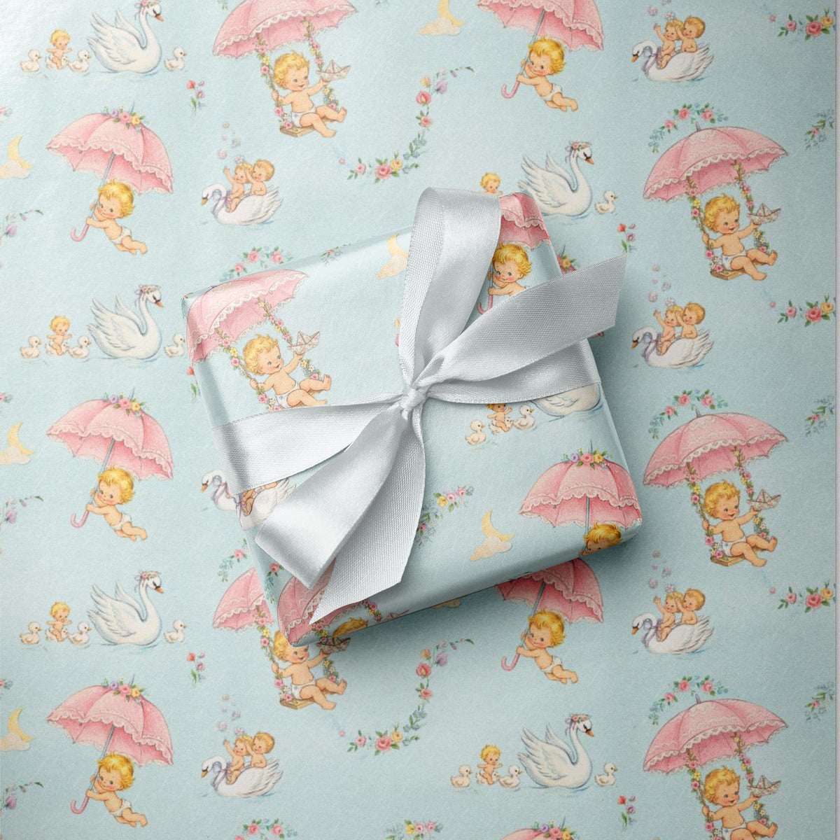 A gift box wrapped in light blue wrapping paper with a pink and blue babies and swan pattern, tied with a white satin ribbon, placed on top of the same wrapping paper.