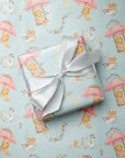 A gift box wrapped in light blue wrapping paper with a pink and blue babies and swan pattern, tied with a white satin ribbon, placed on top of the same wrapping paper.