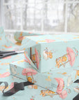 Pastel wrapping paper with a whimsical babies on floral swings pattern, shown wrapped around gift boxes on a table with scissors and ribbon near a bright window.