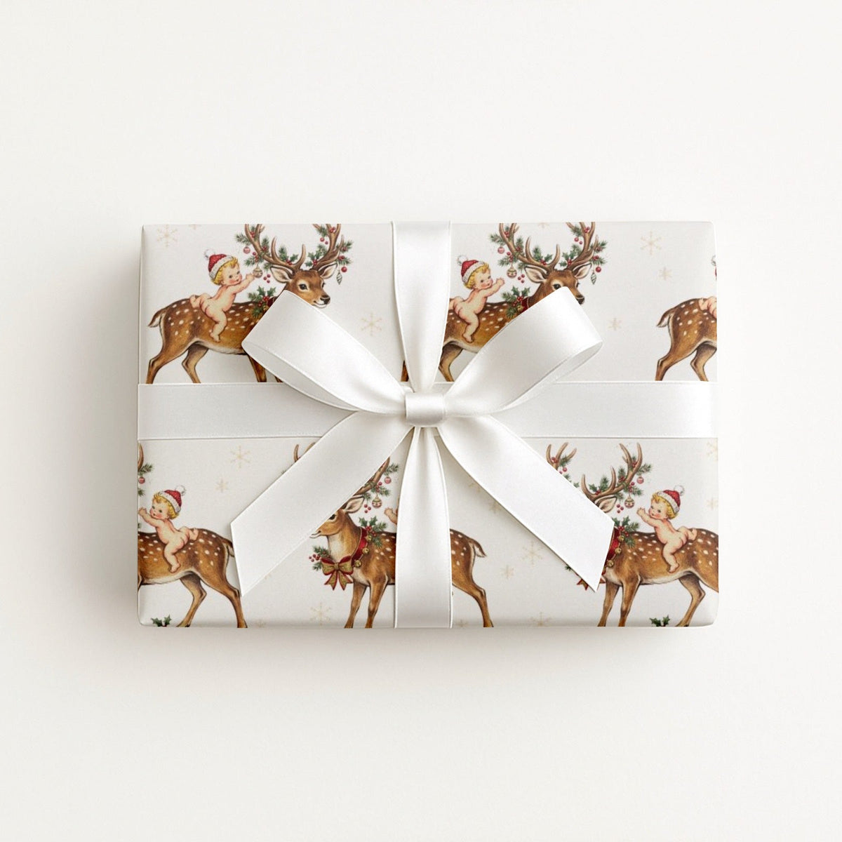 A rectangular gift box wrapped in ivory Christmas-themed paper featuring a repeating pattern of newborn babies riding on festive reindeer, tied with a wide white satin ribbon and bow, on a clean white background.
