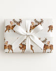 A rectangular gift box wrapped in ivory Christmas-themed paper featuring a repeating pattern of newborn babies riding on festive reindeer, tied with a wide white satin ribbon and bow, on a clean white background.
