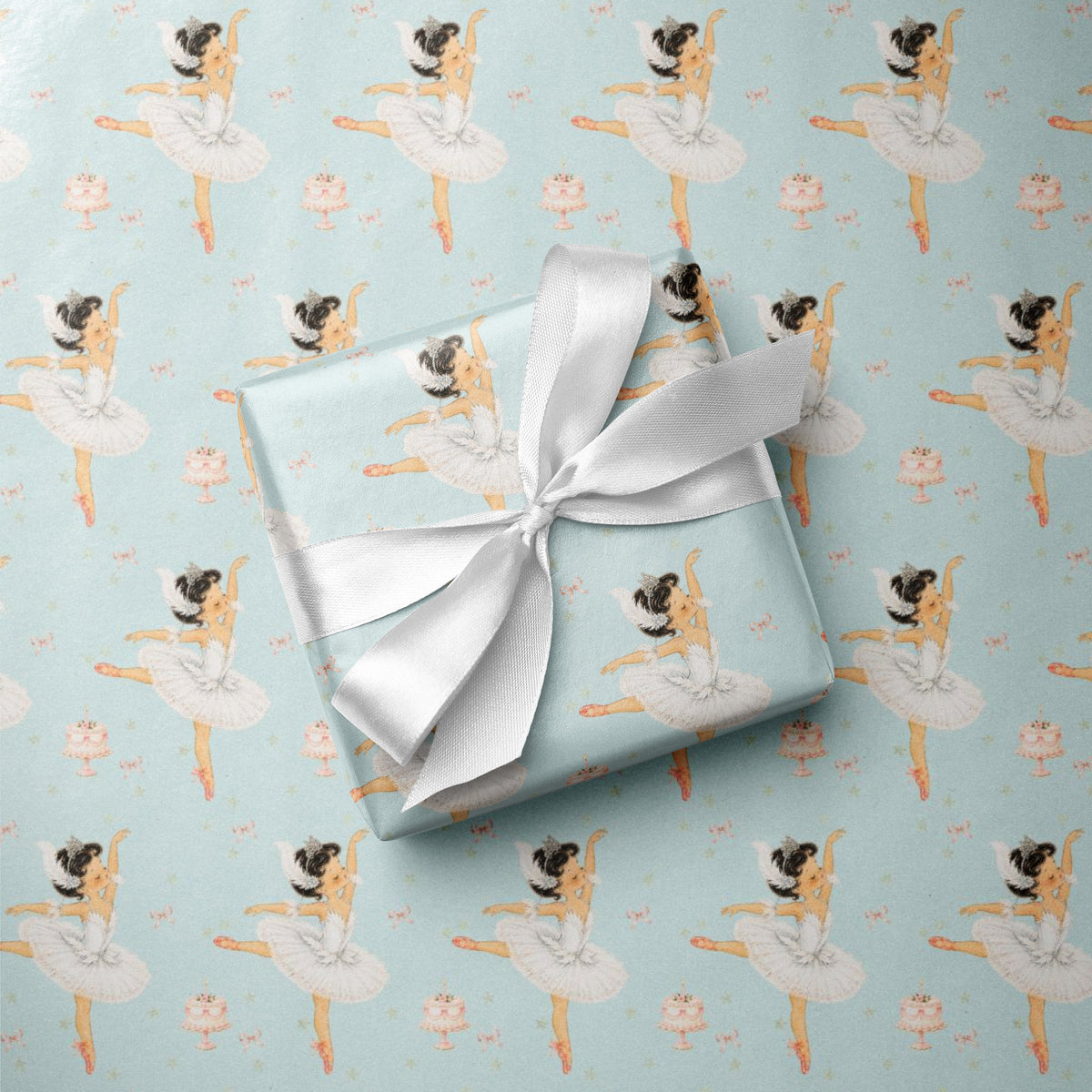 A wrapped gift box with a white satin ribbon sits on top of ballet-themed birthday wrapping paper featuring little girl ballerina illustrations on a light aqua background.