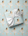 A wrapped gift box with a white satin ribbon sits on top of ballet-themed birthday wrapping paper featuring little girl ballerina illustrations on a light aqua background.