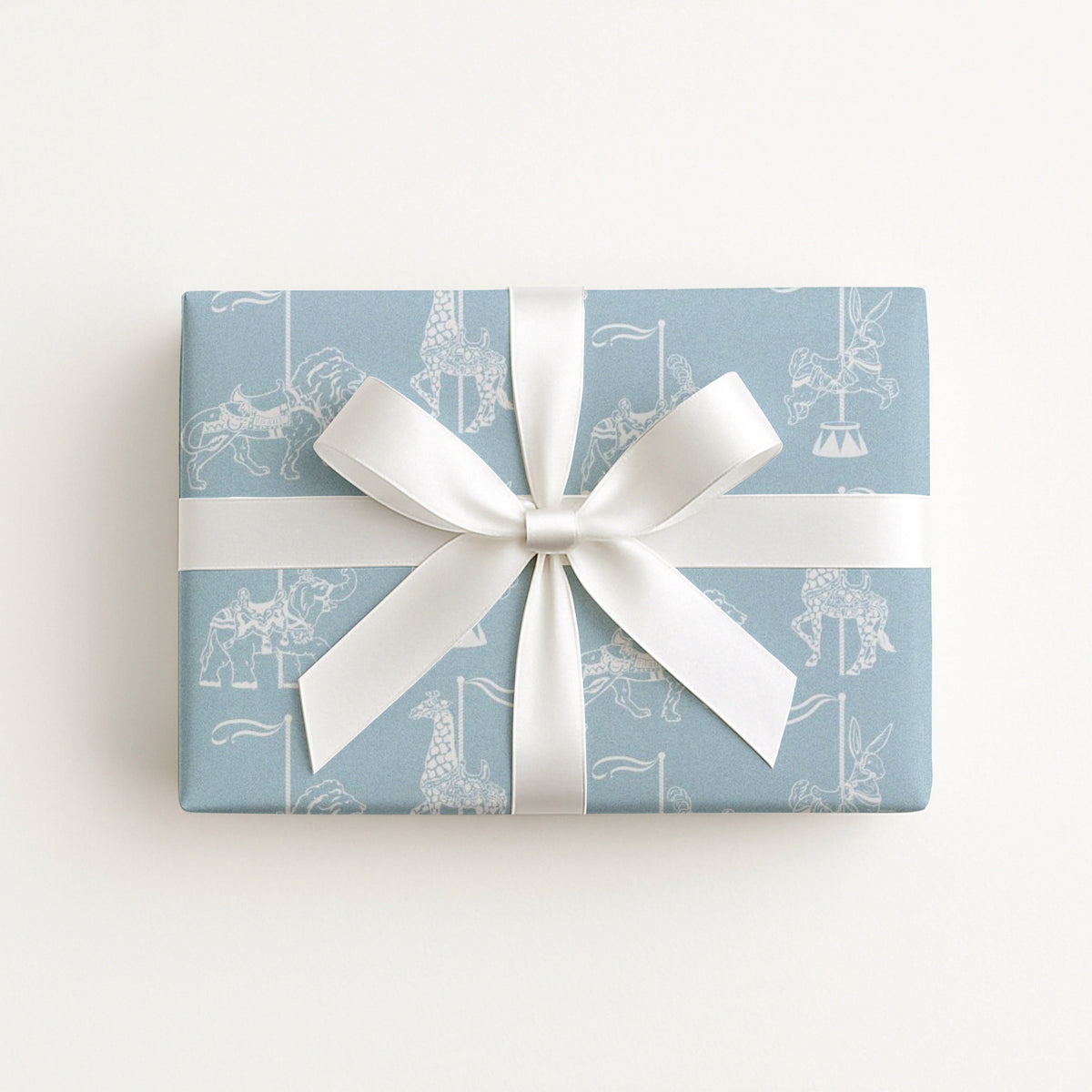 A rectangular gift box wrapped in light blue wrapping paper with a white carousel pattern, tied with a wide white ribbon and bow, on a clean white background.