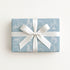 A rectangular gift box wrapped in light blue wrapping paper with a white carousel pattern, tied with a wide white ribbon and bow, on a clean white background.