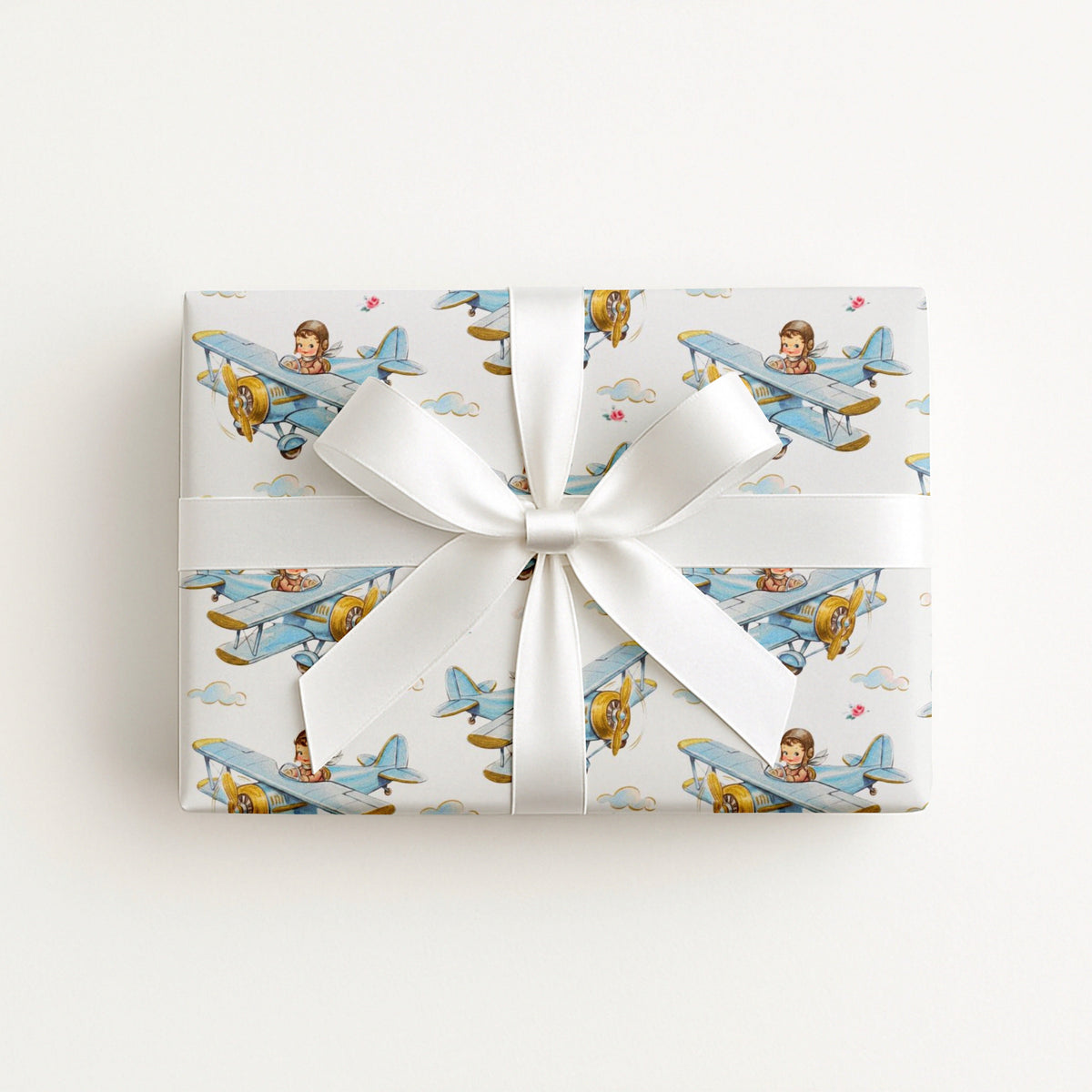 A rectangular gift box wrapped in white paper with a repeating pattern of vintage blue airplanes carrying a boy pilot, tied with a wide white ribbon and bow, on a light off-white background.