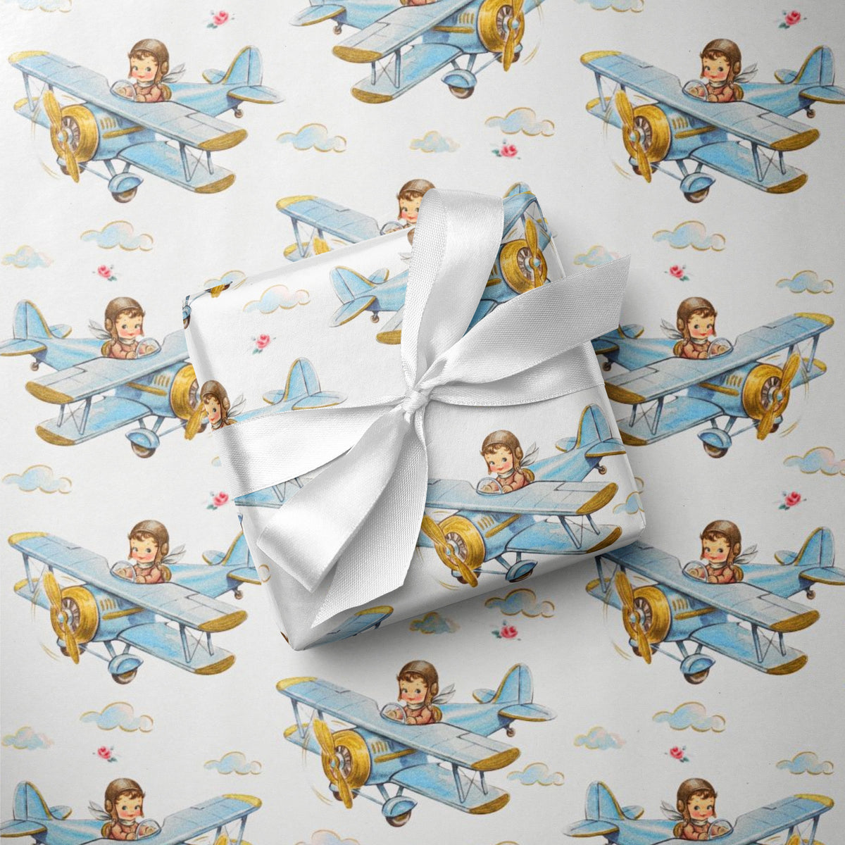 A gift box wrapped in white satin ribbon, placed on top of wrapping paper printed with a repeating pattern of blue biplanes piloted by a baby boy.