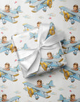 A gift box wrapped in white satin ribbon, placed on top of wrapping paper printed with a repeating pattern of blue biplanes piloted by a baby boy.