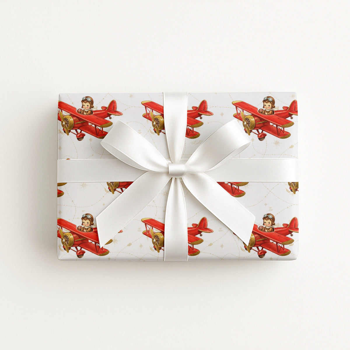 Wrapped gift in vintage-inspired airplane wrapping paper featuring a little boy flying a red biplane over a delicate navigation map with gold accents on an ivory background, tied with a white satin bow.