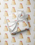 Gift wrapped in elegant wedding cake wrapping paper with a white satin bow, resting on a sheet of the repeating tiered cake pattern