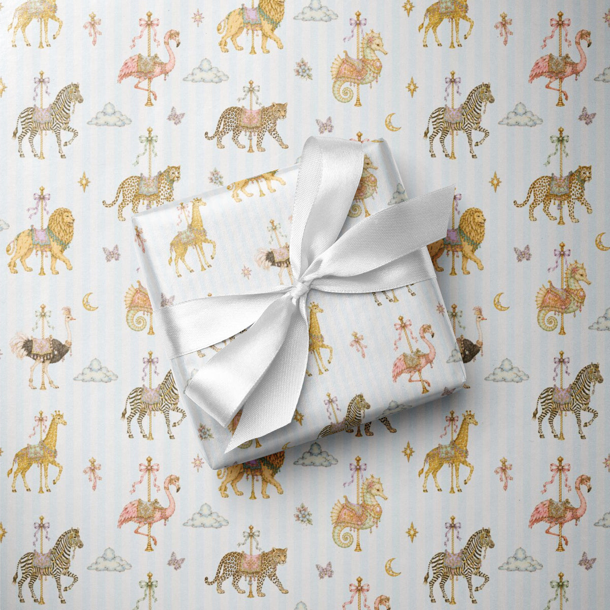 A wrapped gift box tied with a white satin ribbon, placed on top of pale blue wrapping paper printed with a whimsical carousel and animal pattern in soft gold, pink, and brown tones.