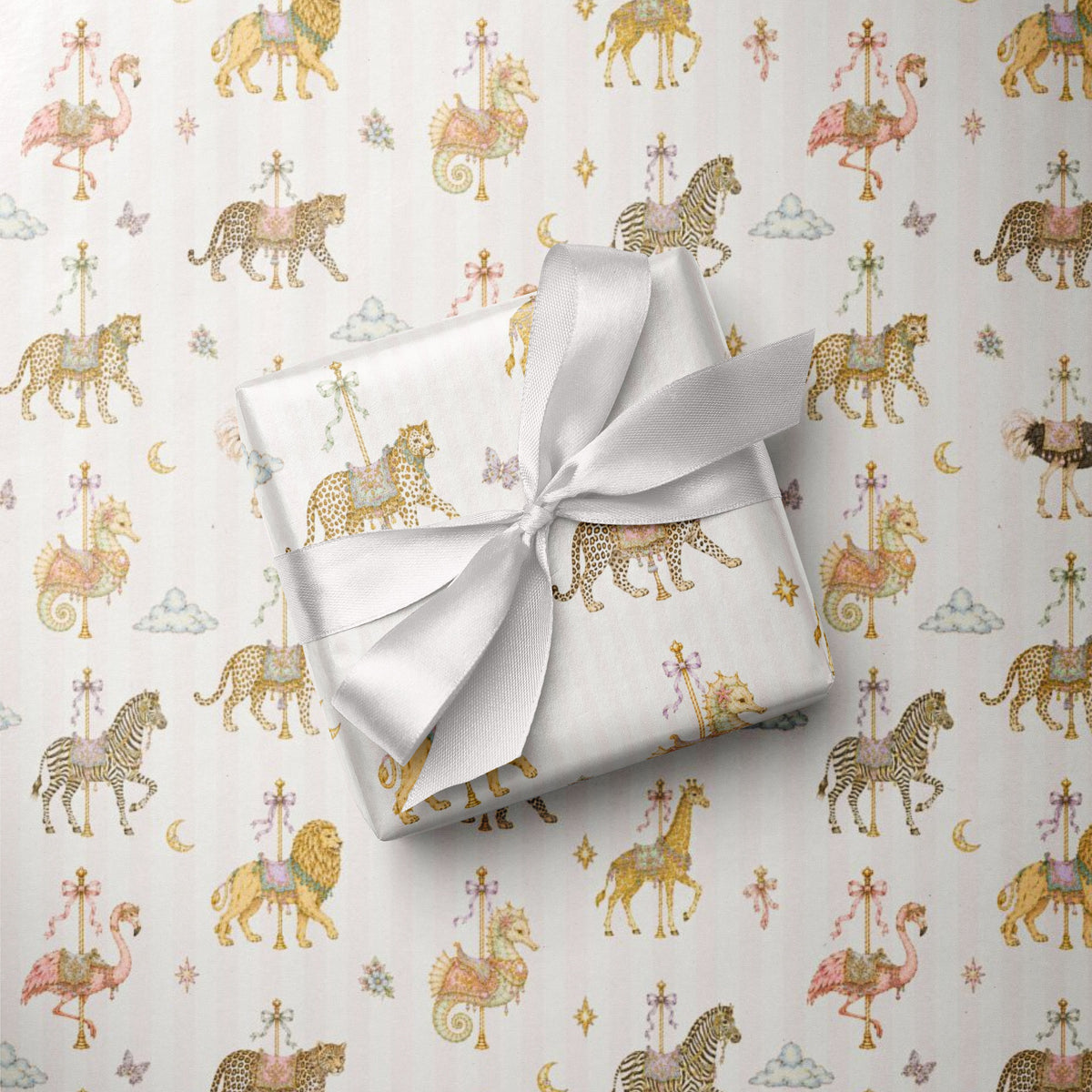 A wrapped gift box tied with a white satin ribbon, placed on top of pale pink wrapping paper printed with a whimsical carousel and animal pattern in soft gold, pink, and brown tones.