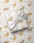 A wrapped gift box tied with a white satin ribbon, placed on top of pale pink wrapping paper printed with a whimsical carousel and animal pattern in soft gold, pink, and brown tones.