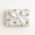 A rectangular gift box wrapped in light blue wrapping paper with a carousel and animal print, tied with a wide white satin ribbon and bow, photographed from above on a clean white background.