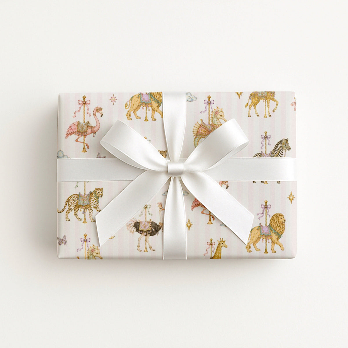A rectangular gift box wrapped in pale pink wrapping paper with a whimsical carousel print featuring lions, giraffes, and zebra-like animals, tied with a wide white satin ribbon and bow, on a clean white background.