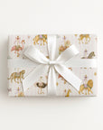 A rectangular gift box wrapped in pale pink wrapping paper with a whimsical carousel print featuring lions, giraffes, and zebra-like animals, tied with a wide white satin ribbon and bow, on a clean white background.