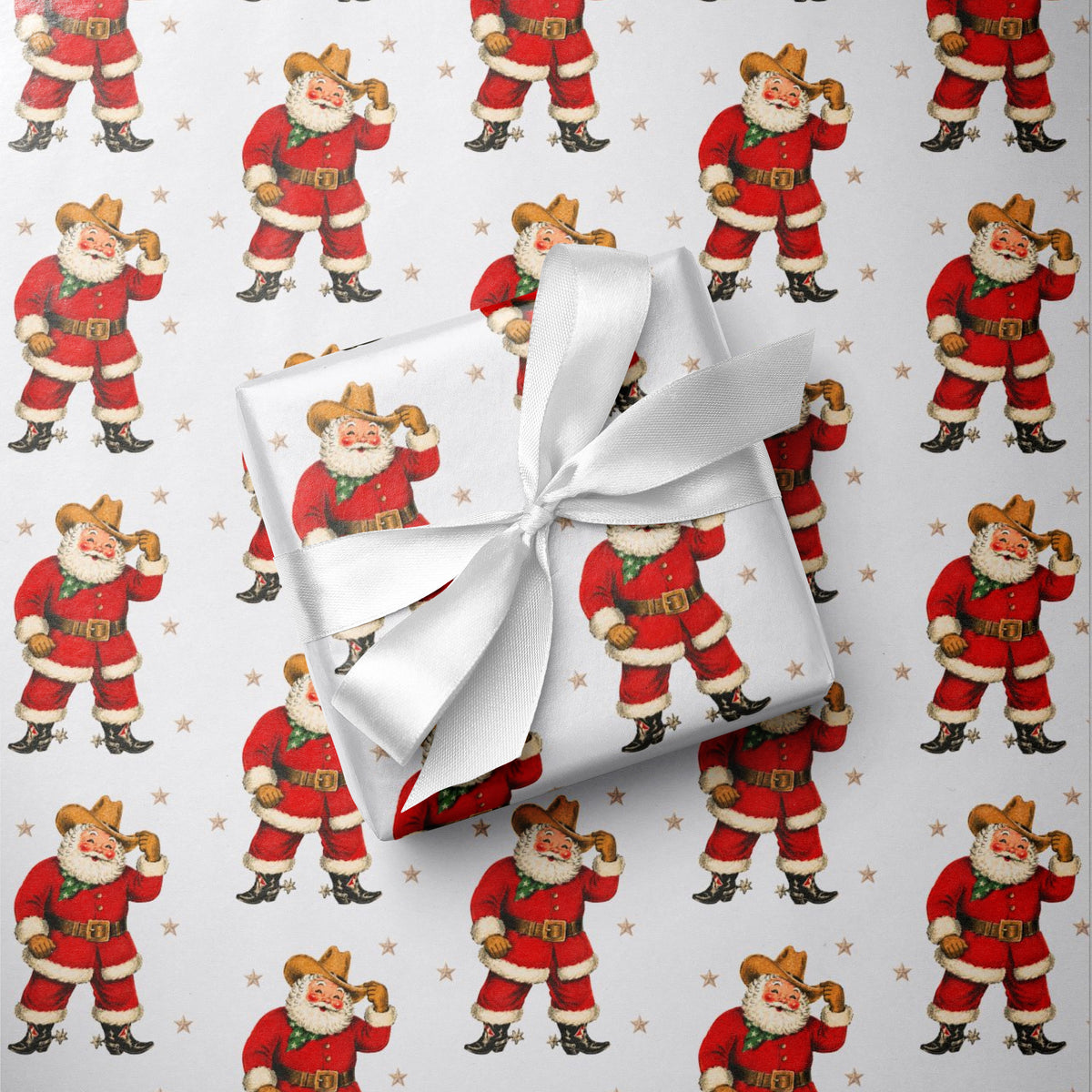 A gift box wrapped in white paper with a large white ribbon bow, placed on top of Christmas-themed wrapping paper featuring a repeating pattern of western Santa Claus in a cowboy outfit.