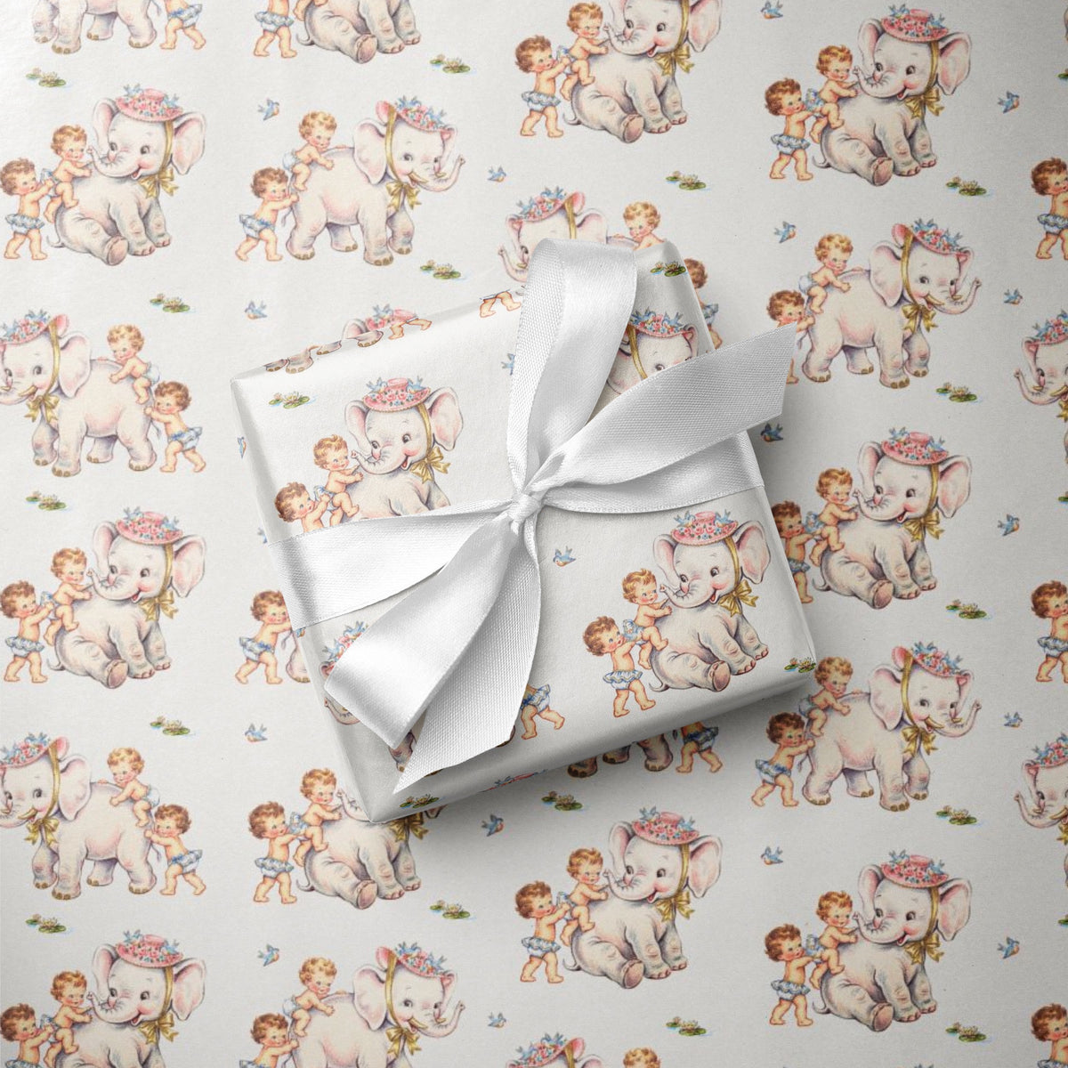 A gift box wrapped in ivory paper with a repeating pattern of vintage-style elephants and children, tied with a white satin ribbon, placed on top of the same patterned wrapping paper.