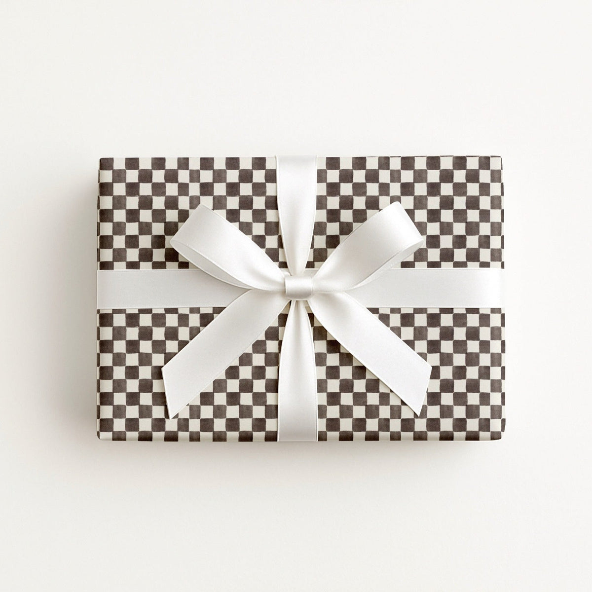 A rectangular gift box wrapped in brown and white checkered patterned paper, tied with a wide white satin ribbon and bow, photographed from above on a clean white background.