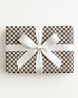 A rectangular gift box wrapped in brown and white checkered patterned paper, tied with a wide white satin ribbon and bow, photographed from above on a clean white background.