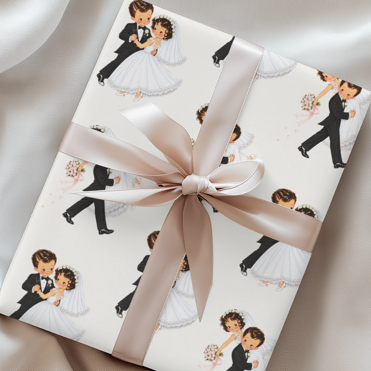 A gift box wrapped in white paper printed with a repeating illustration of a dancing bride and groom, tied with a wide satin ribbon in a large bow, set against a soft white fabric background.
