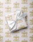 A wrapped baby gift tied with a white satin ribbon, placed on top of matching gift wrap printed with a repeating vintage carousel pattern on a pale pink background.