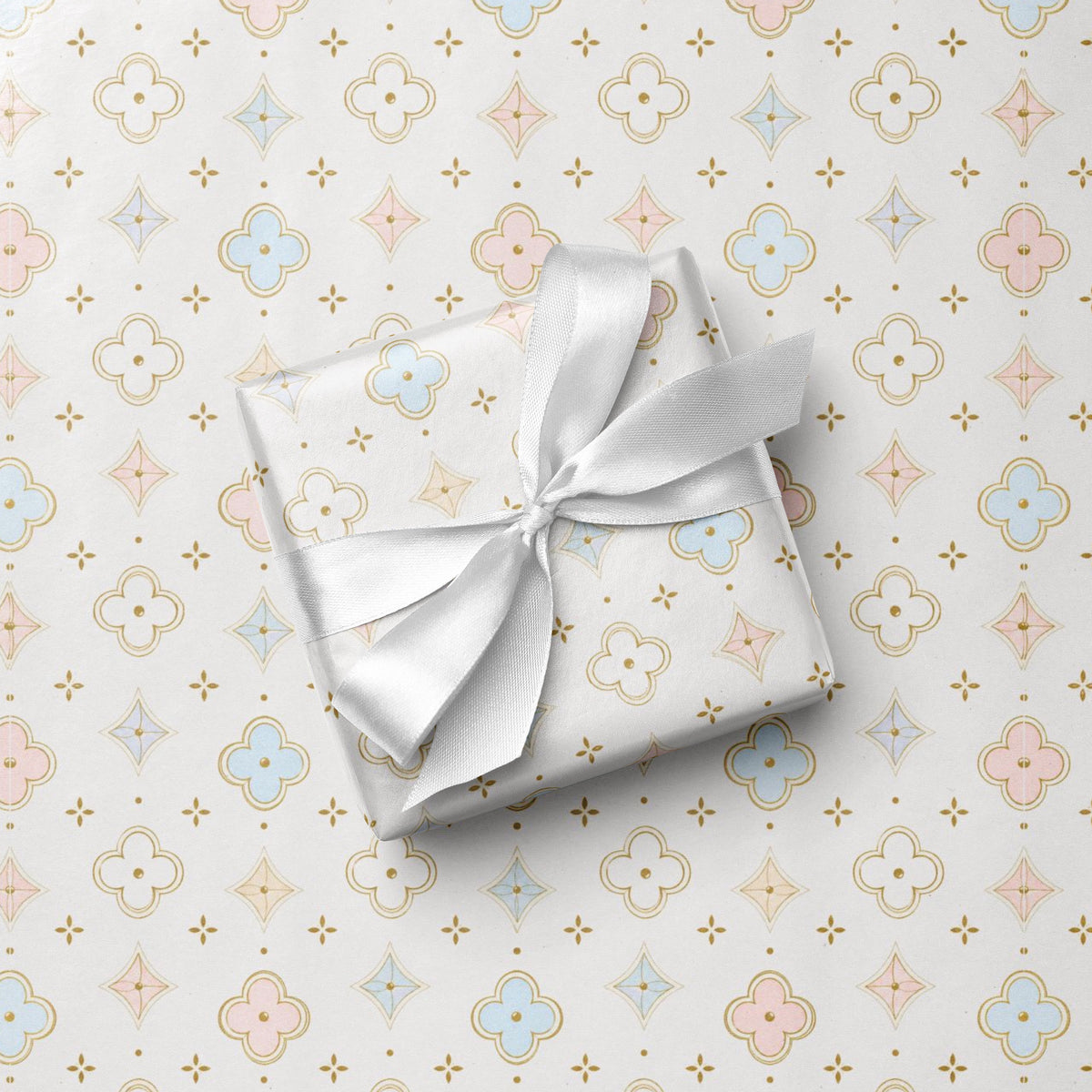A square gift box wrapped in ivory paper with a matching white satin ribbon tied in a bow, placed on top of patterned wrapping paper featuring a repeating quatrefoil and star motif in soft pink, blue, and gold.