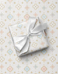 A square gift box wrapped in ivory paper with a matching white satin ribbon tied in a bow, placed on top of patterned wrapping paper featuring a repeating quatrefoil and star motif in soft pink, blue, and gold.