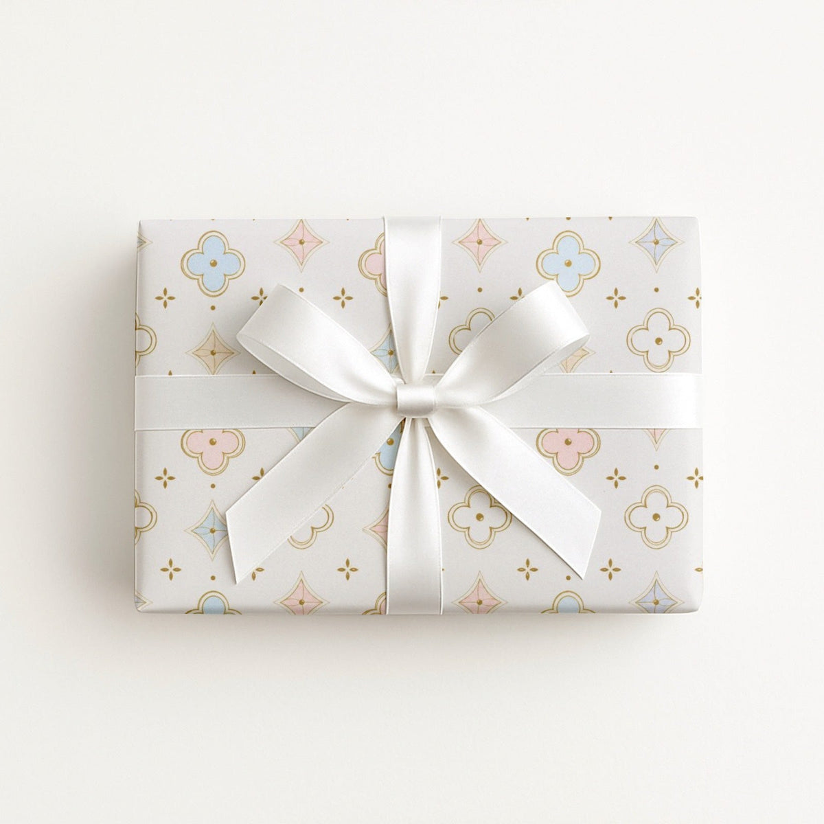 A rectangular gift box wrapped in white paper with a delicate pastel pattern of gold, pink, and blue motifs, tied with a large white satin ribbon in a bow, set against a clean white background.