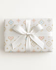 A rectangular gift box wrapped in white paper with a delicate pastel pattern of gold, pink, and blue motifs, tied with a large white satin ribbon in a bow, set against a clean white background.