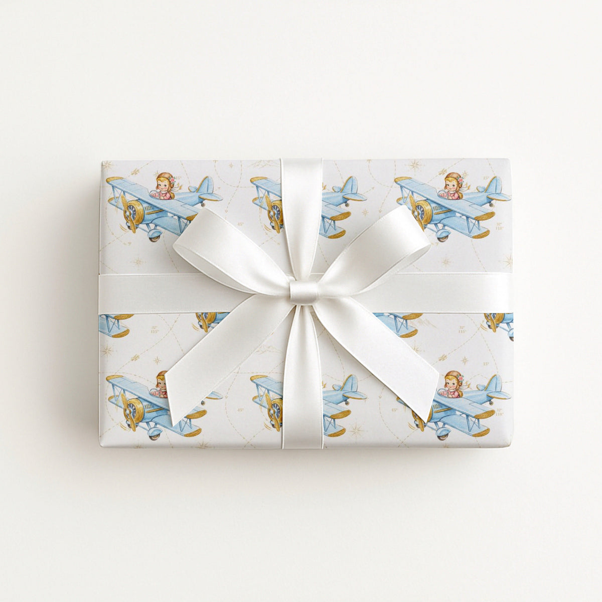 Wrapped gift in vintage-inspired airplane wrapping paper featuring a little girl flying a powder blue biplane over a delicate navigation map with gold accents on an ivory background, tied with a white satin bow.