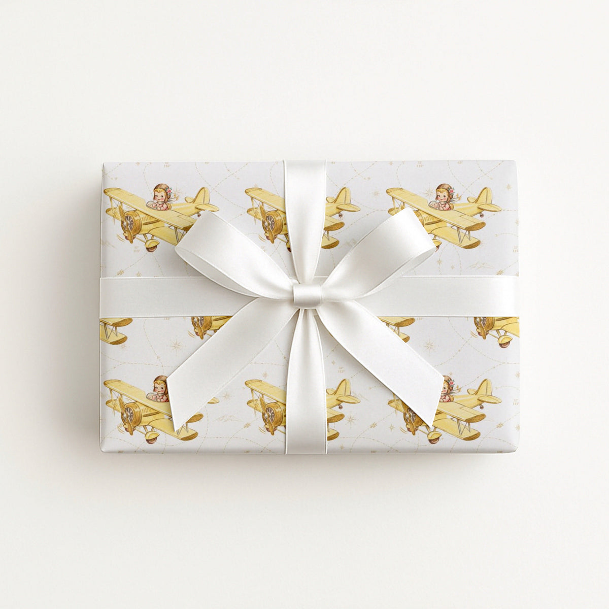 Wrapped gift in vintage-inspired airplane wrapping paper featuring a little girl flying a yellow and gold biplane over a delicate navigation map with gold accents on an ivory background, tied with a white satin bow.
