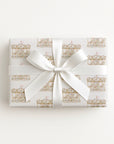 A rectangular gift box wrapped in white paper with a repeating gold carousel pattern and tied with a wide white satin ribbon bow, centered on a plain white background.