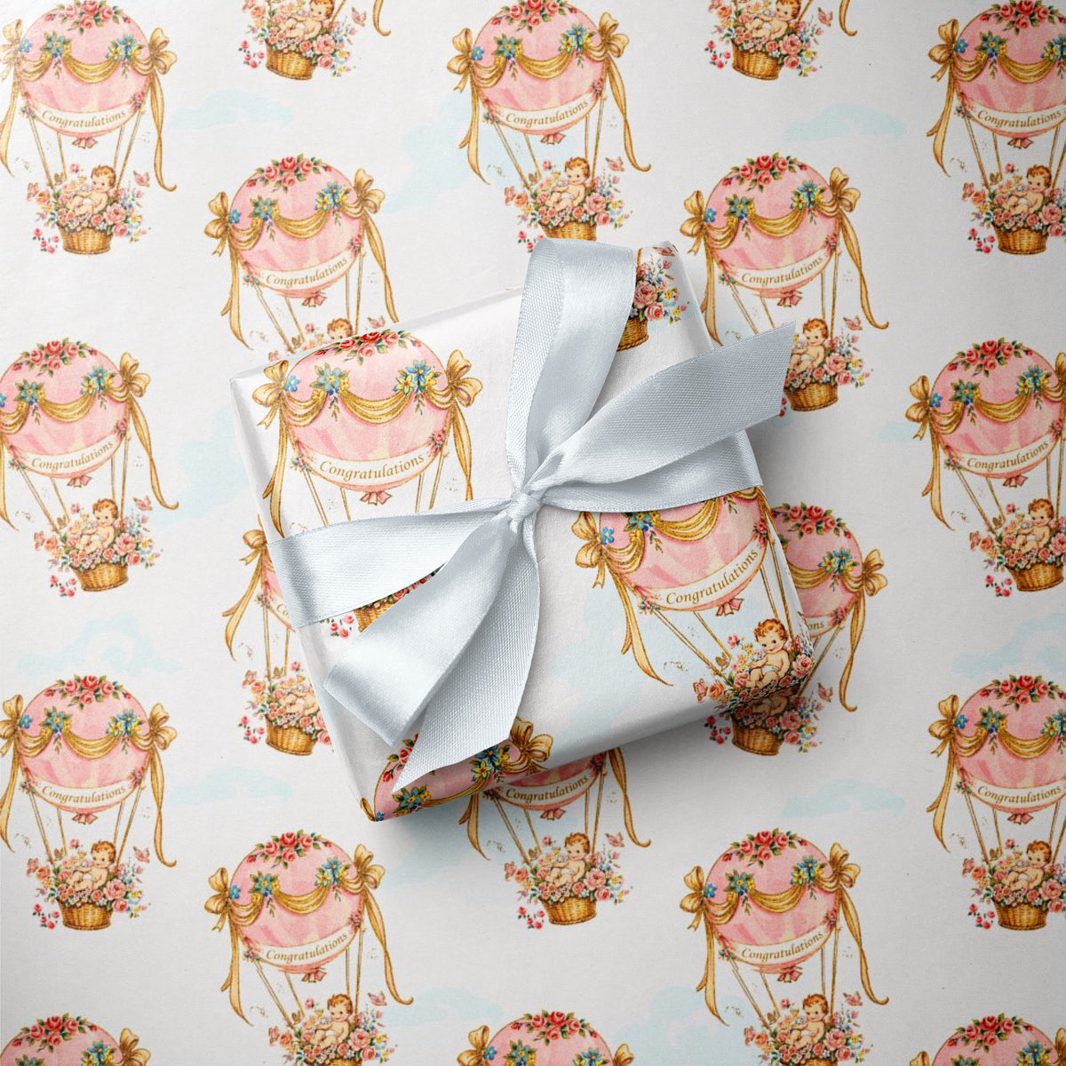 A gift box wrapped in pastel pink and cream wrapping paper with a repeating hot air balloon pattern, tied with a white satin ribbon, placed on top of matching wrapping paper.
