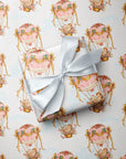 A gift box wrapped in pastel pink and cream wrapping paper with a repeating hot air balloon pattern, tied with a white satin ribbon, placed on top of matching wrapping paper.