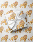 A gift wrapped in white satin ribbon with a bow, placed on top of matching wrapping paper printed with a repeating pattern of golden lions, flower crowns and sweet babies.