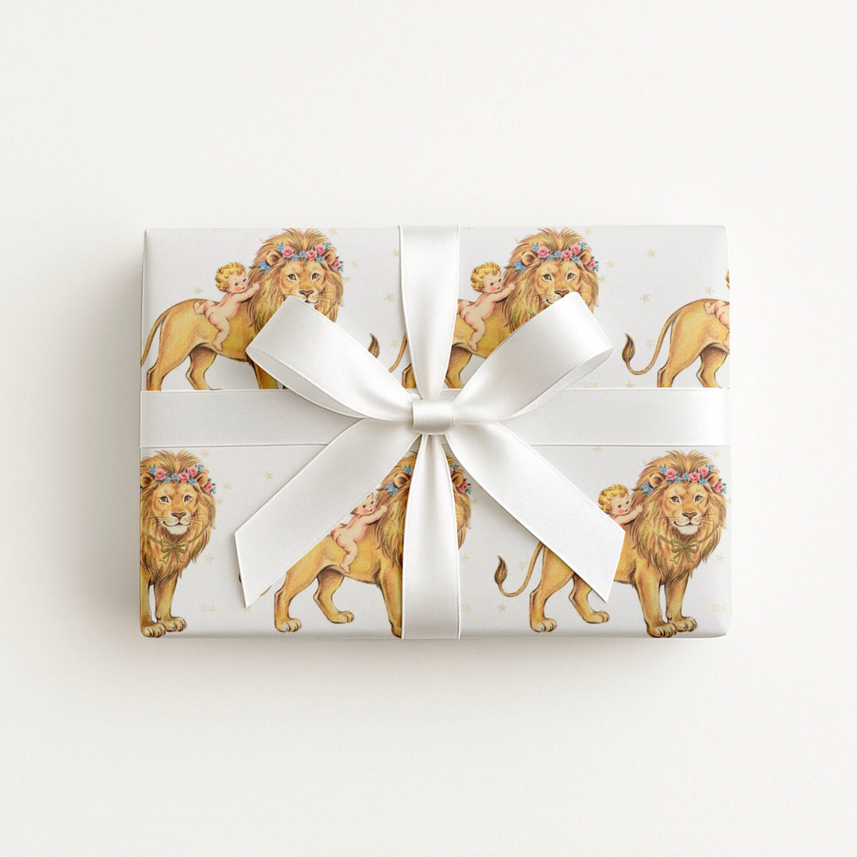 A rectangular gift box wrapped in white paper featuring a repeating pattern of golden lions in floral crowns and babies, tied with a large white satin bow on a clean white background.