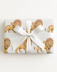 A rectangular gift box wrapped in white paper featuring a repeating pattern of golden lions in floral crowns and babies, tied with a large white satin bow on a clean white background.